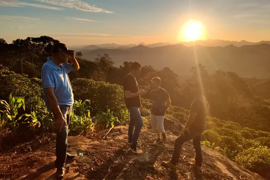 Vier Männer stehen auf einem Hügel bei Sonnenaufgang. Im Hintergrund sind Berge zu sehen.