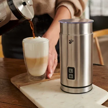 Frau gießt Kaffee in ein Glas mit Milch neben einem elektrischen Milchaufschäumer aus Edelstahl.