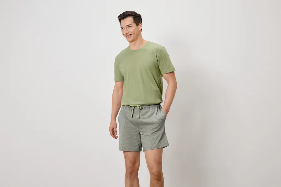 Mann mit olivgrünem T-Shirt und grauen Shorts mit Kordelzug, die Hände in den Hosentaschen, lächelnd.