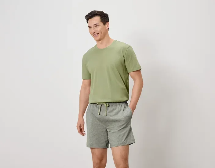 Mann mit olivgrünem T-Shirt und grauen Shorts mit Kordelzug, die Hände in den Hosentaschen, lächelnd.