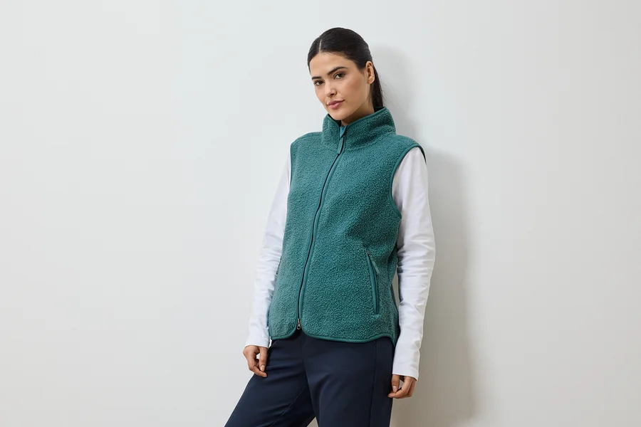 Frau mit Pferdeschwanz, die eine grüne Fleeceweste über einem weißen Langarmshirt und einer marineblauen Hose trägt.