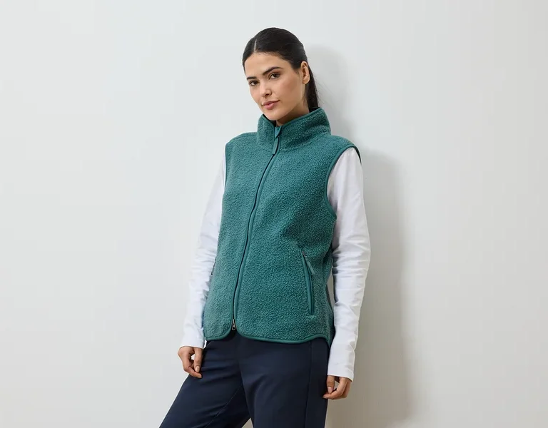 Frau mit Pferdeschwanz, die eine grüne Fleeceweste über einem weißen Langarmshirt und einer marineblauen Hose trägt.