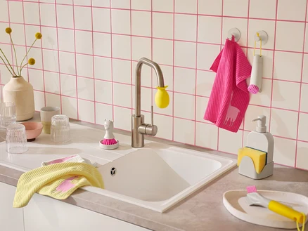 Eine Küchenspüle mit weißer Fliesenwand und rosa Fugen. Auf der Arbeitsplatte liegen gelbe und pinke Geschirrtücher mit Lama-Motiven, Spülbürsten und ein Seifenspender mit Schwammhalter.