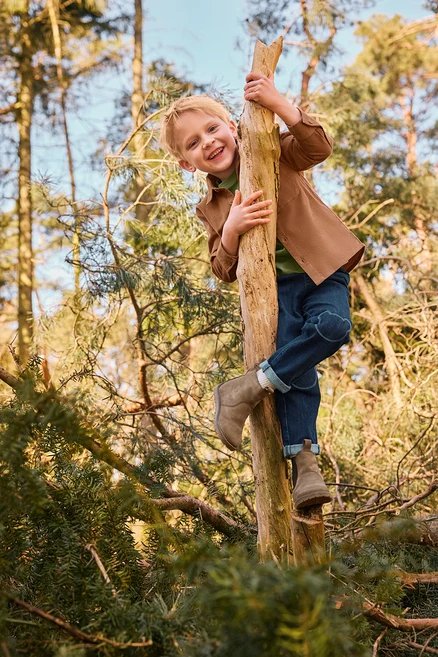 Ein lächelnder kleiner Junge klettert in einem Wald an einem Baumstamm hoch. Er trägt eine braune Jacke, ein grünes Shirt, blaue Jeans mit Knie-Patches und hellbraune Stiefel.