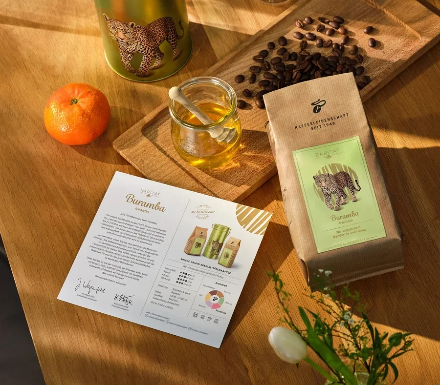 Auf einem Holztisch liegen eine Dose, eine Packung und ein Zettel Kaffee, eine Tasse Kaffee, eine Mandarine, Honig und eine Vase mit Tulpen.