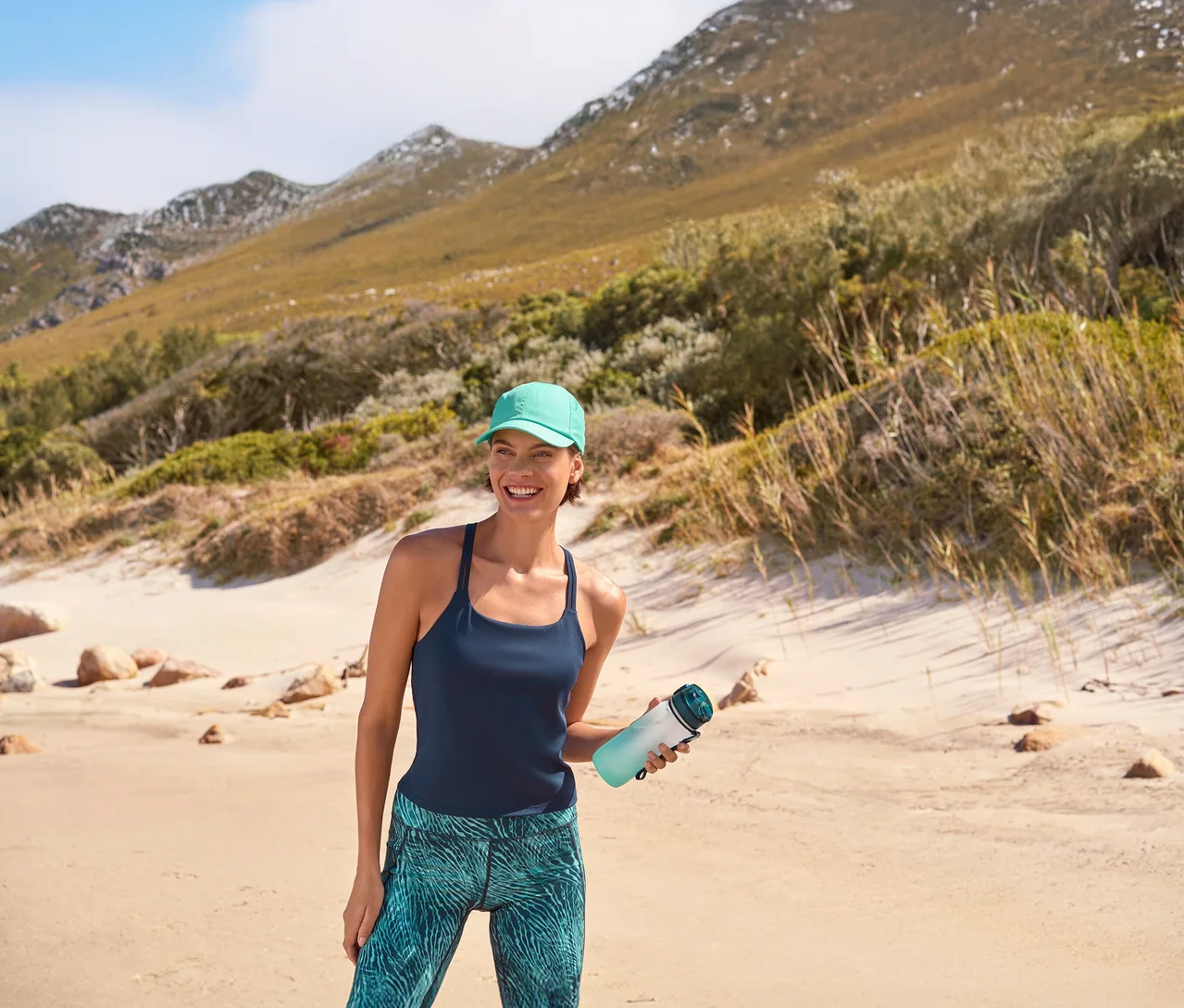 Lächelnde Frau in türkisener Sport-Kappe, dunkelblauem Yogatop mit integriertem BH und Sportradler »Active Power« mit Alloverprint, die eine Trinkflasche mit Kühlakku am Strand hält. Im Hintergrund sind Berge zu sehen.