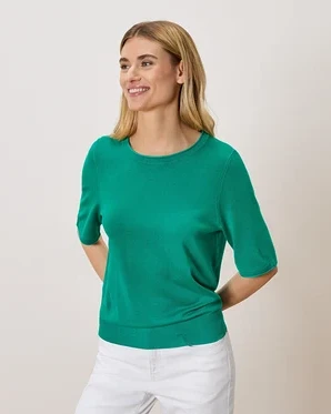 Frau mit blonden Haaren, die einen grünen Pullover und weiße Hosen trägt.