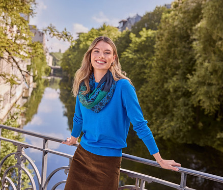 Lächelnde Frau in blauem Oberteil, braunem Rock und gemustertem Schal, trägt Doppel-Creolen aus 925 Silber rhodiniert, lehnt an einer Brücke über einem Fluss.
