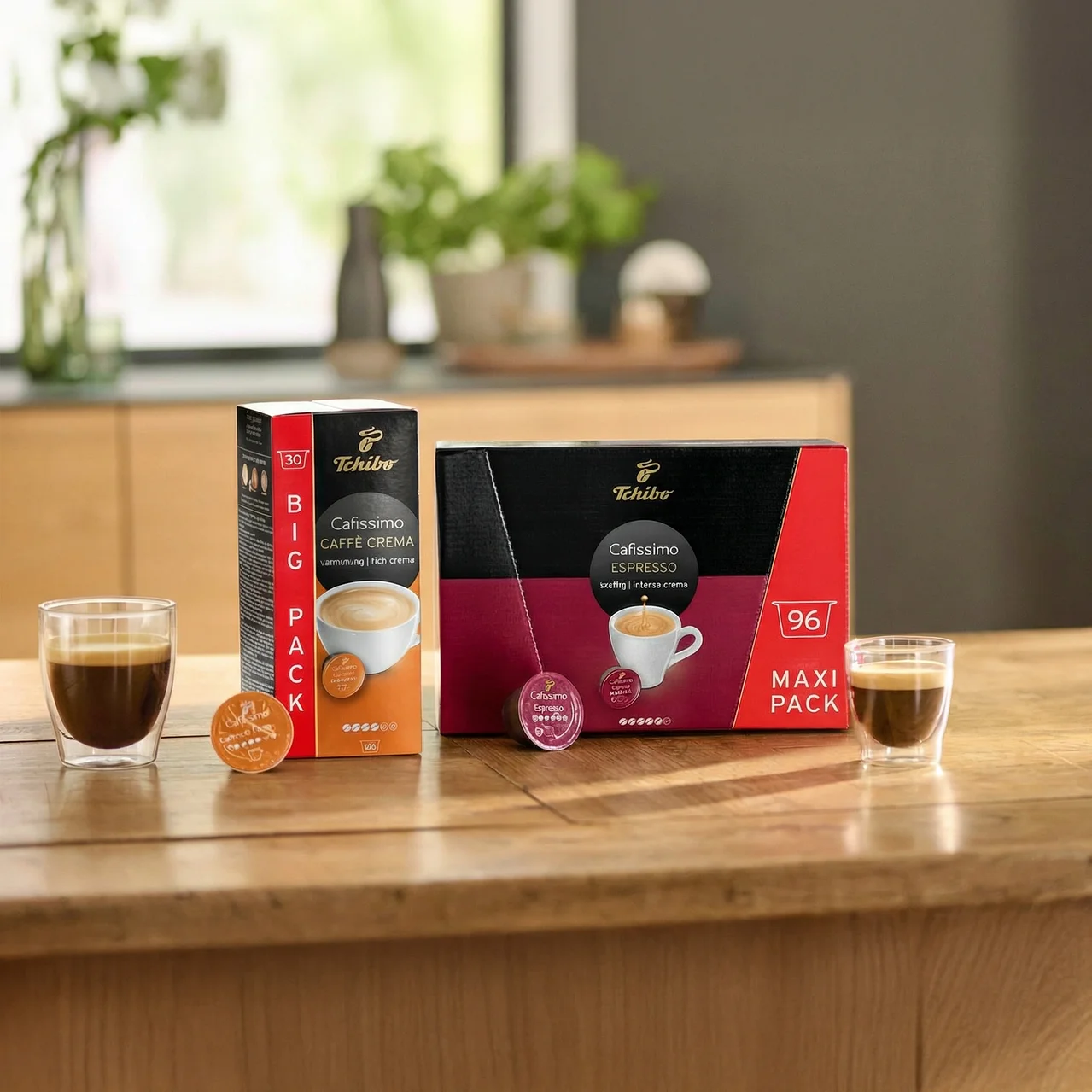 Auf einem Holztisch stehen zwei Gläser Espresso neben Packungen Tchibo Cafissimo Caffe Crema und Espresso.