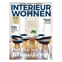 Magazincover für 'INTERIEUR & WOHNEN' zeigt ein modernes Esszimmer mit rundem Glastisch und vier Stühlen mit blauen Polstern und goldenen, skulpturalen Füßen. Im Hintergrund sind große Fenster sichtbar. Der Titel 'Wohnen mit Klassikern' ist aufgedruckt.