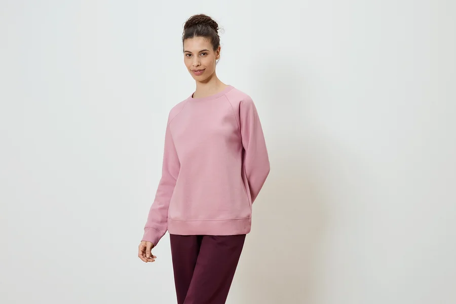 Frau trägt ein rosafarbenes Sweatshirt und eine burgunderfarbene Hose vor einem weißen Hintergrund.