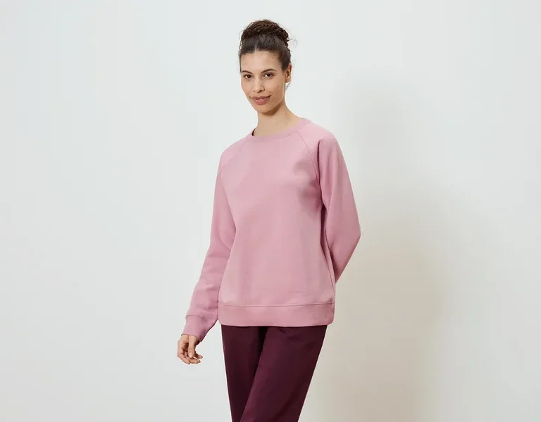 Frau trägt ein rosafarbenes Sweatshirt und eine burgunderfarbene Hose vor einem weißen Hintergrund.