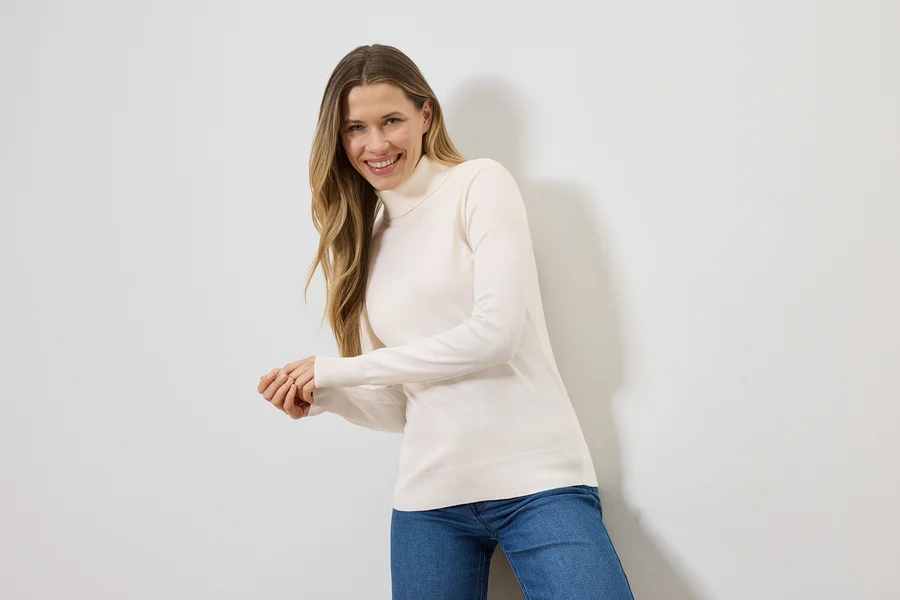Frau mit blonden Haaren lächelt in einem cremefarbenen Rollkragenpullover und blauen Jeans vor einer weißen Wand.