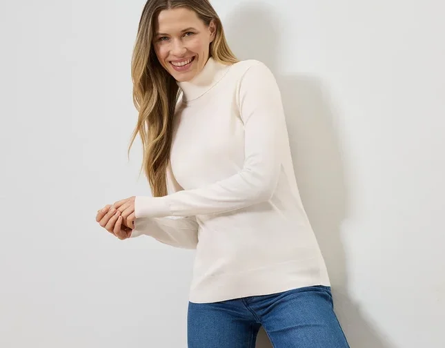 Frau mit blonden Haaren lächelt in einem cremefarbenen Rollkragenpullover und blauen Jeans vor einer weißen Wand.