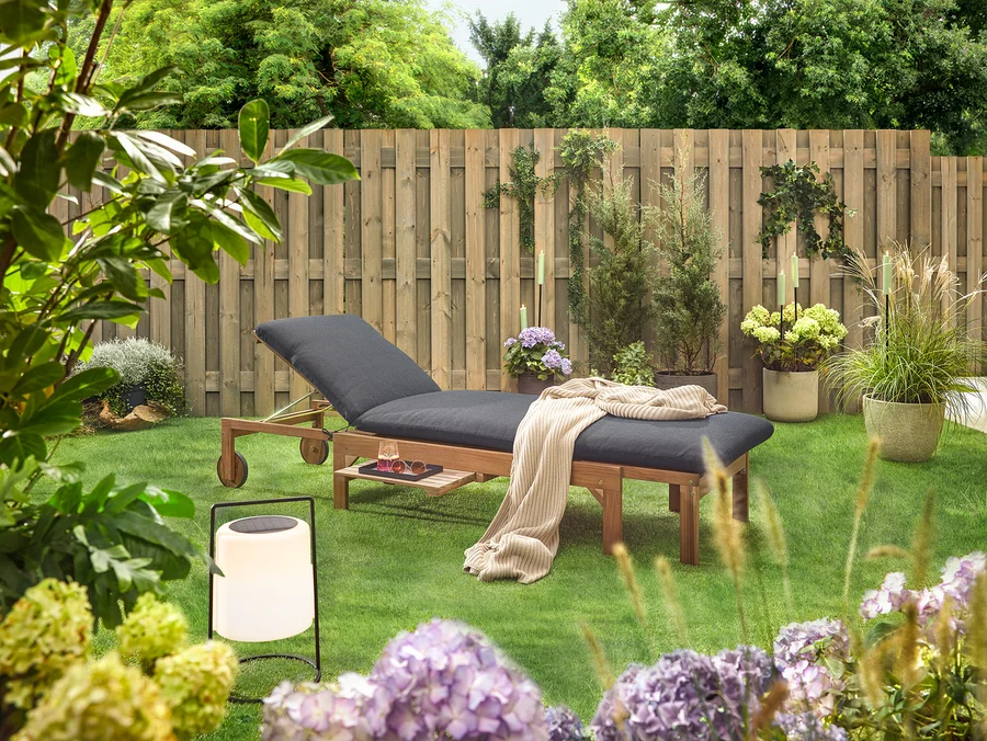 Gartenszene mit einer hölzernen Liege mit schwarzer Polsterung, einer Decke, einem Getränk, einer Sonnenbrille und einer Laterne auf einem Rasen vor einem Holzzaun.