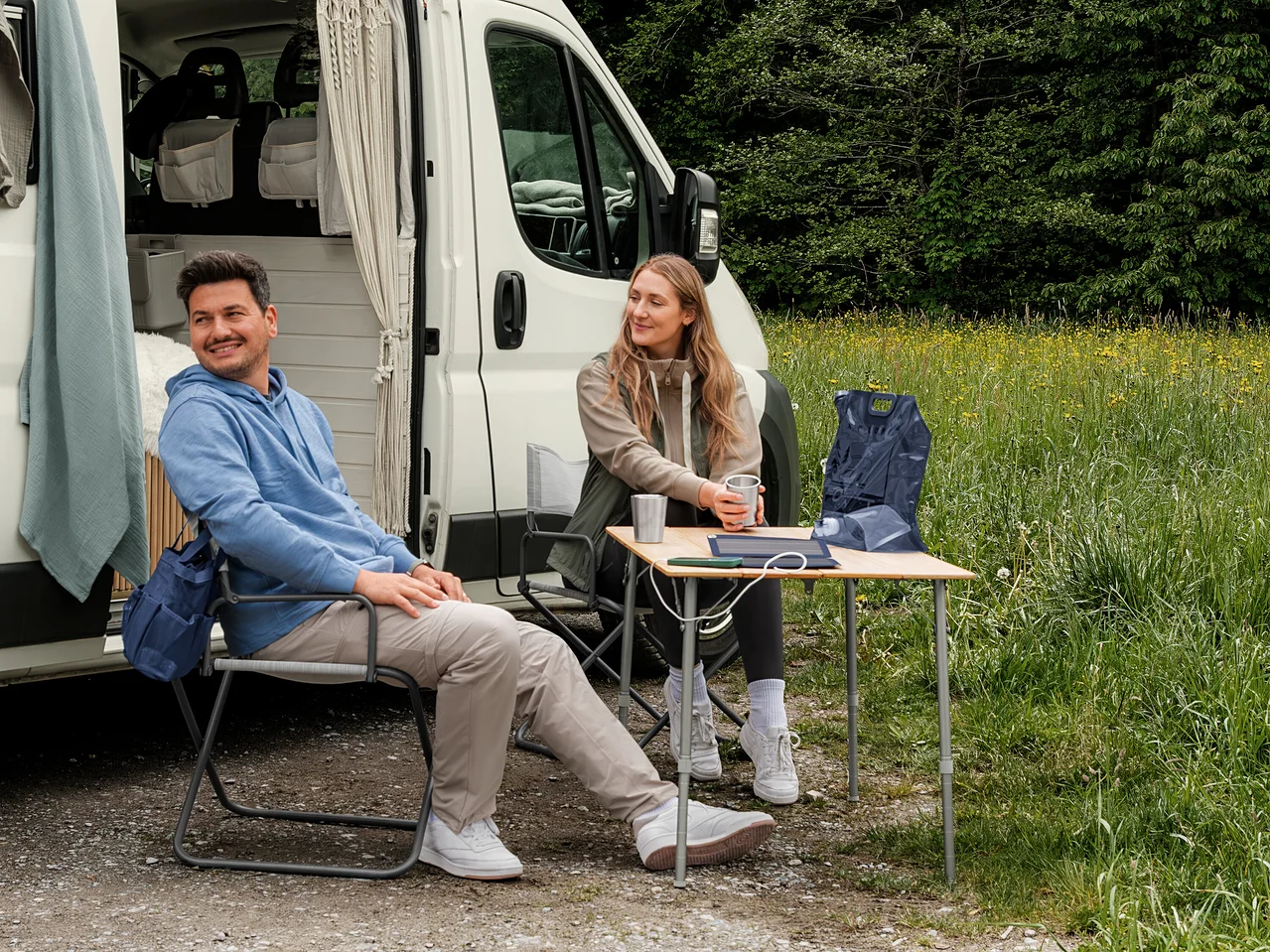 Ein Mann in einem hellblauen Sweat-Hoodie und eine Frau in einer beigen Sweatjacke und einer khakifarbenen Hybrid-Weste sitzen vor einem Campervan. Auf einem Tisch liegt eine dunkelblaue Pack-Weste.