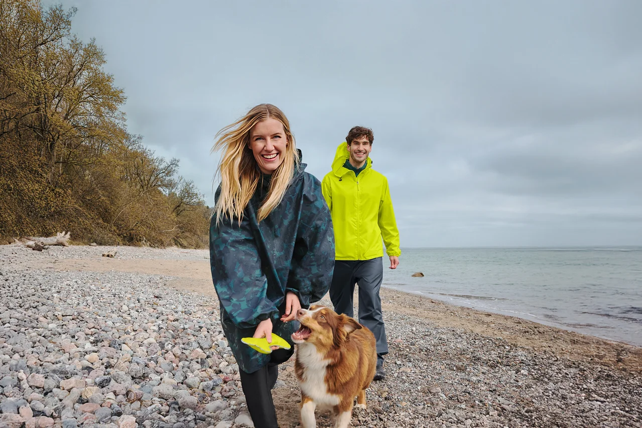 Eine lächelnde Frau in einem dunklen Regenponcho hält einen gelben Frisbee, während sie mit einem braun-weißen Hund an einem Kiesstrand spaziert. Ein lächelnder Mann in einer gelben Regenjacke geht hinter ihnen, mit Meer und bewölktem Himmel im Hintergrund.