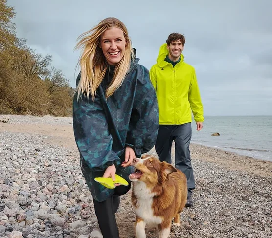 Eine lächelnde Frau in einem dunklen Regenponcho hält einen gelben Frisbee, während sie mit einem braun-weißen Hund an einem Kiesstrand spaziert. Ein lächelnder Mann in einer gelben Regenjacke geht hinter ihnen, mit Meer und bewölktem Himmel im Hintergrund.