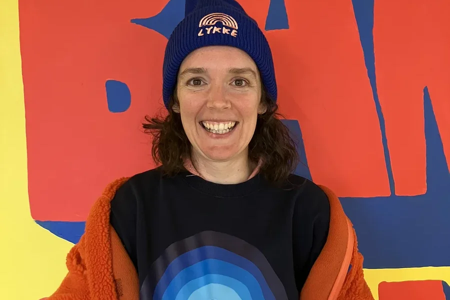 Eine lächelnde Frau mit dunklen Haaren blickt in die Kamera. Sie trägt eine blaue Mütze mit „LYKKE“-Schriftzug und Regenbogen, ein dunkelblaues Sweatshirt mit blauem Regenbogenmotiv und eine orangefarbene flauschige Jacke über den Schultern. Der Hintergrund ist ein farbenfrohes Wandgemälde mit großen roten Formen auf gelbem und blauem Grund.