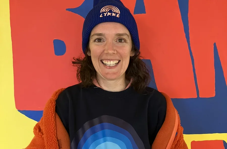 Eine lächelnde Frau mit dunklen Haaren blickt in die Kamera. Sie trägt eine blaue Mütze mit „LYKKE“-Schriftzug und Regenbogen, ein dunkelblaues Sweatshirt mit blauem Regenbogenmotiv und eine orangefarbene flauschige Jacke über den Schultern. Der Hintergrund ist ein farbenfrohes Wandgemälde mit großen roten Formen auf gelbem und blauem Grund.