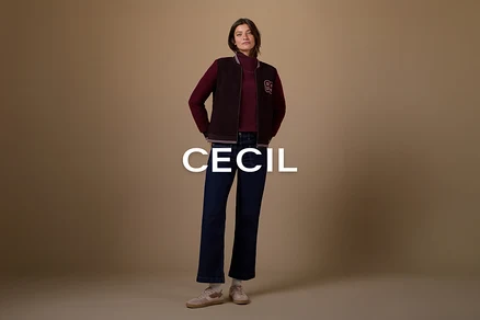 Eine Frau steht mit den Händen in den Taschen und trägt einen dunkelroten Rollkragenpullover, eine dunkelbraune Bomberjacke mit einem weißen „B“-Aufnäher, dunkelblaue Schlagjeans und hellrosa Sneaker mit weißen Socken vor einem einfarbigen braunen Hintergrund.
