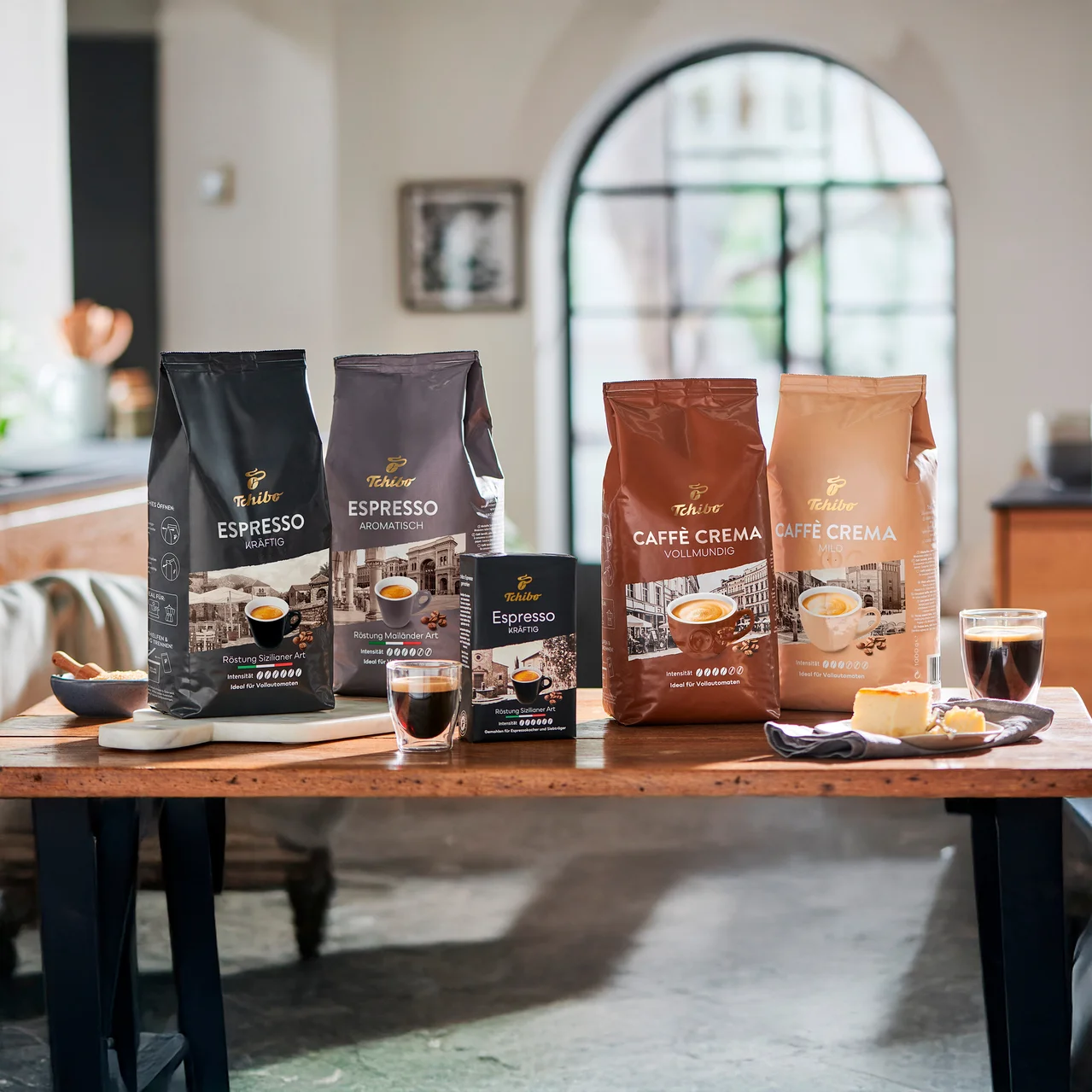 Vier Packungen Tchibo Espresso und Caffè Crema Kaffee auf einem Holztisch mit einer Tasse Kaffee und einem Stück Kuchen.