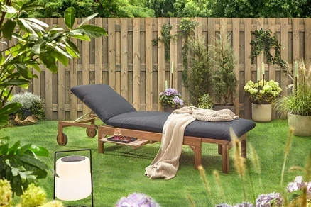 Gartenszene mit einer hölzernen Liege mit schwarzer Polsterung, einer Decke, einem Getränk, einer Sonnenbrille und einer Laterne auf einem Rasen vor einem Holzzaun.