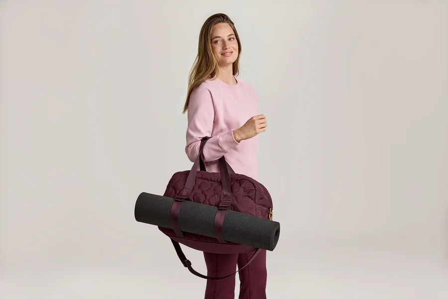 Frau mit rosa Pullover und weinroter Hose, die eine weinrote Sporttasche mit einer schwarzen Yogamatte über der Schulter trägt.