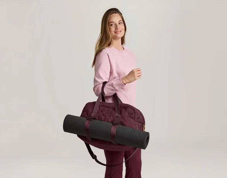 Frau mit rosa Pullover und weinroter Hose, die eine weinrote Sporttasche mit einer schwarzen Yogamatte über der Schulter trägt.
