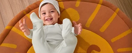 Ein lächelndes Baby liegt auf einem Kinderzimmer-Teppich »Löwe« und trägt ein 3-teiliges Baby-Set.