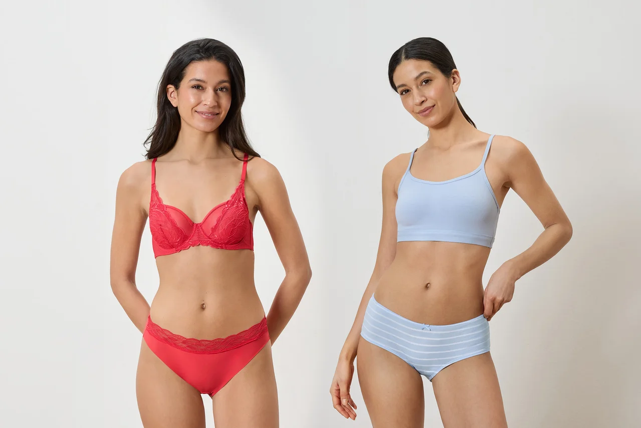 Zwei Frauen stehen nebeneinander. Die linke Frau trägt einen roten Spitzen-BH und passende rote Slips mit Spitzenbesatz. Die rechte Frau trägt einen hellblauen Bralette und hellblaue gestreifte Boxershorts.