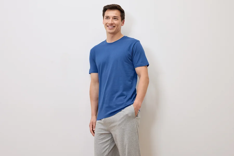 Lächelnder Mann in blauem T-Shirt und grauer Jogginghose posiert vor weißem Hintergrund.