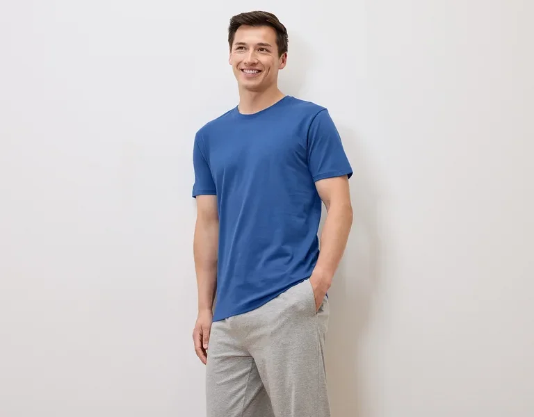 Lächelnder Mann in blauem T-Shirt und grauer Jogginghose posiert vor weißem Hintergrund.