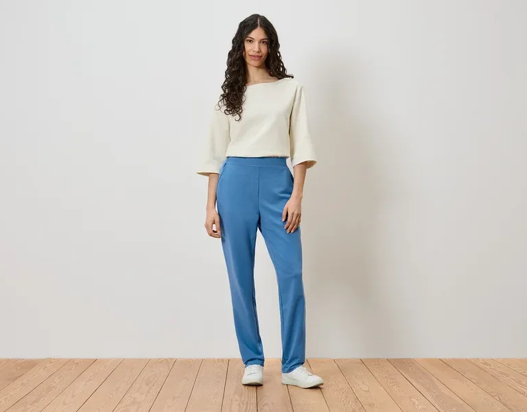 Eine Frau steht vor einer Wand und trägt ein cremefarbenes Oberteil, blaue Hosen und weiße Schuhe.