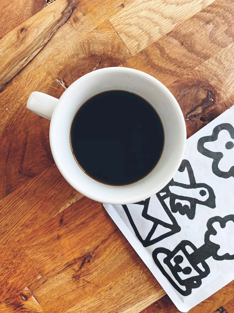 Draufsicht auf eine Tasse Kaffee neben einer weißen Serviette mit schwarzen Zeichnungen auf einem Holztisch.