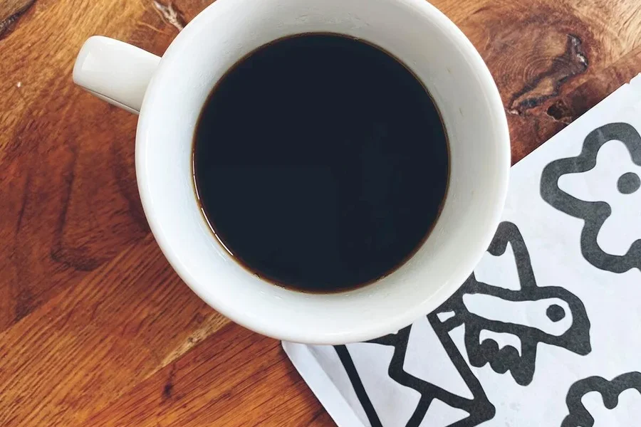 Draufsicht auf eine Tasse Kaffee neben einer weißen Serviette mit schwarzen Zeichnungen auf einem Holztisch.