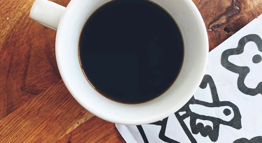 Draufsicht auf eine Tasse Kaffee neben einer weißen Serviette mit schwarzen Zeichnungen auf einem Holztisch.
