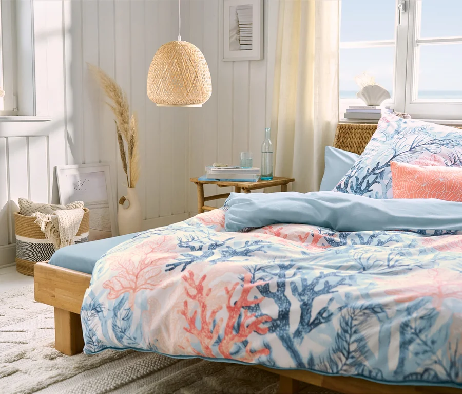 Schlafzimmer mit einem Bett, hellblaues Microfaser-Spannbettlaken in Doppelgröße, Renforcé-Bettwäsche und zwei hellblaue und zwei korallenfarbene Microfaser-Kopfkissenbezüge. Über dem Bett hängt eine Pendelleuchte.