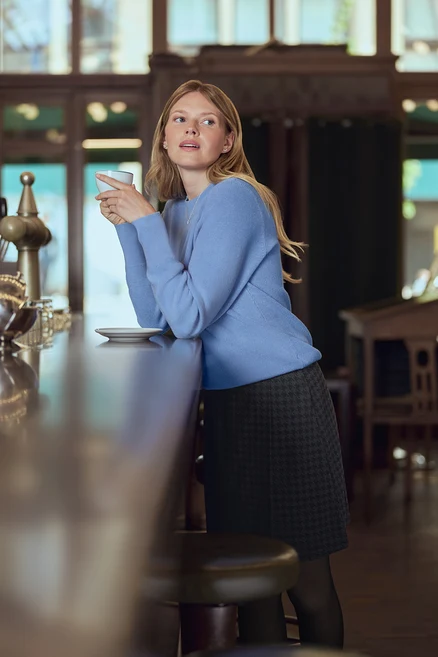 Frau im blauen Pullover hält eine Tasse an einem Tresen. Sie trägt 925 Silber Ohrstecker Zirkoniabogen.