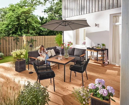 Auf einer Terrasse sitzt eine Frau mit einem Hund auf einer Dining-Lounge-Ecke »Elin«. Ein Grillwagen mit Polyzement-Arbeitsfläche, ein Pflanztopf mit Textilgeflecht, eine LED-Akku-Outdoor-Stehleuchte, eine LED-Akku-Outdoor-Tischleuchte, zwei Outdoor-Kissen mit Leinen-Haptik und ein Gartenstuhl mit Textil-Kreuzgeflecht sind ebenfalls zu sehen.