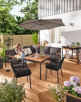 Auf einer Terrasse sitzt eine Frau mit einem Hund auf einer Dining-Lounge-Ecke »Elin«. Ein Grillwagen mit Polyzement-Arbeitsfläche, ein Pflanztopf mit Textilgeflecht, eine LED-Akku-Outdoor-Stehleuchte, eine LED-Akku-Outdoor-Tischleuchte, zwei Outdoor-Kissen mit Leinen-Haptik und ein Gartenstuhl mit Textil-Kreuzgeflecht sind ebenfalls zu sehen.