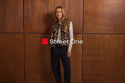 Eine Frau mit langen blonden Haaren steht vor einer Holzwand und trägt eine Weste mit Leopardenmuster über einem weißen Langarmshirt und dunklen Jeans. Sie hat die Hände in den Taschen. Das Logo "Street One" ist sichtbar.