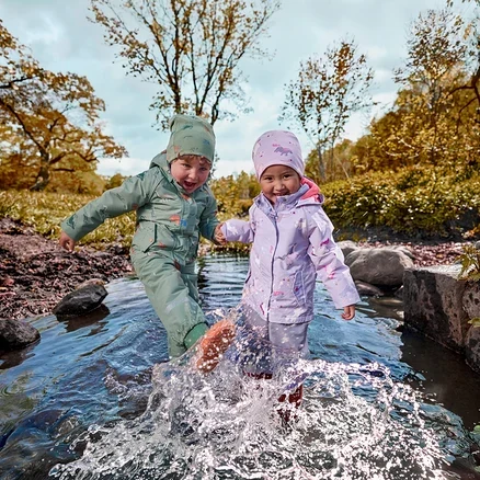 Zwei kleine Kinder in grünen und lila wasserdichten Anzügen planschen im flachen Bach, halten Händchen, mit Herbstbäumen im Hintergrund.