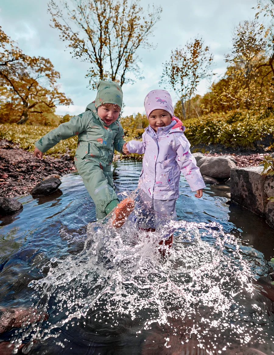 Zwei kleine Kinder in grünen und lila wasserdichten Anzügen planschen im flachen Bach, halten Händchen, mit Herbstbäumen im Hintergrund.