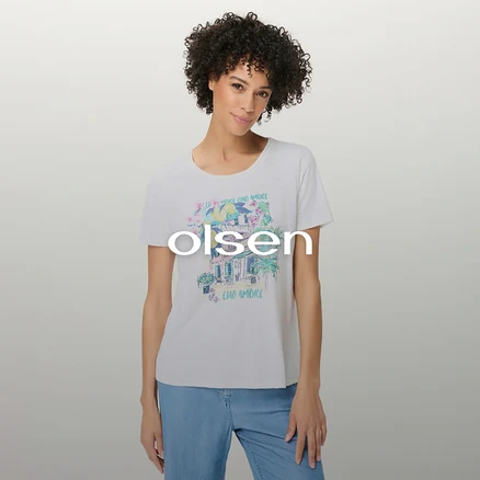 Frau mit lockigem Haar trägt ein weißes T-Shirt mit farbigem Aufdruck eines Gebäudes, Blumen, einer Palme und dem Schriftzug „CIAO AMORE“, dazu hellblaue Jeans.