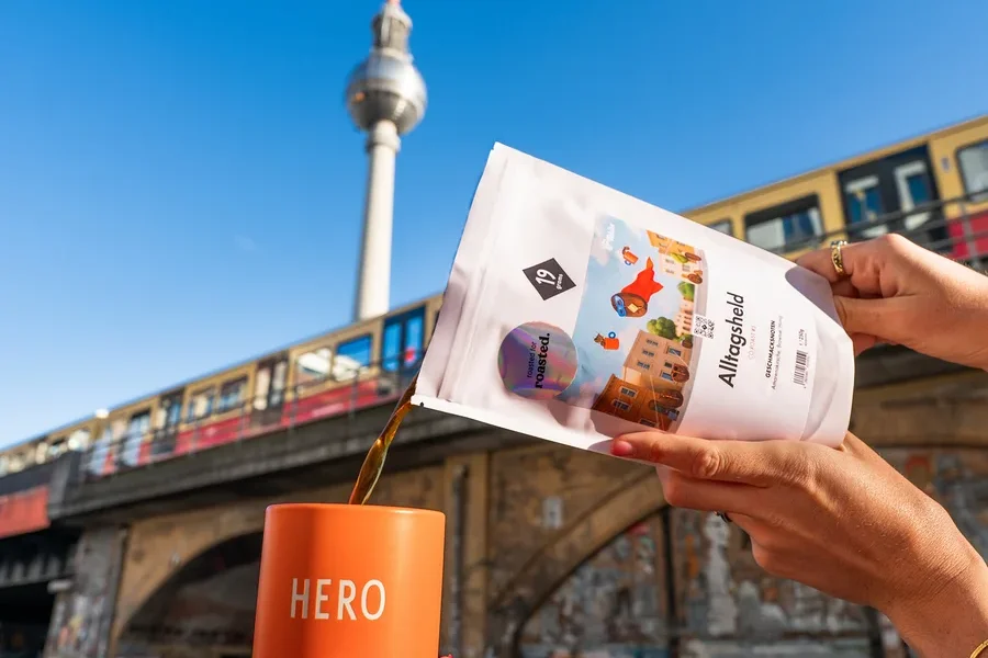 Eine Person gießt Kaffee aus einer weißen Packung in eine orangefarbene Tasse mit der Aufschrift HERO, im Hintergrund der Berliner Fernsehturm.