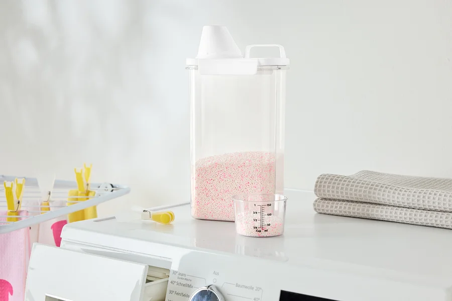 Auf einer Waschmaschine stehen ein durchsichtiger Behälter mit rosa Waschmittel, ein Messbecher und gefaltete Handtücher.