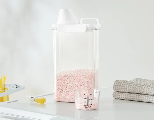 Auf einer Waschmaschine stehen ein durchsichtiger Behälter mit rosa Waschmittel, ein Messbecher und gefaltete Handtücher.