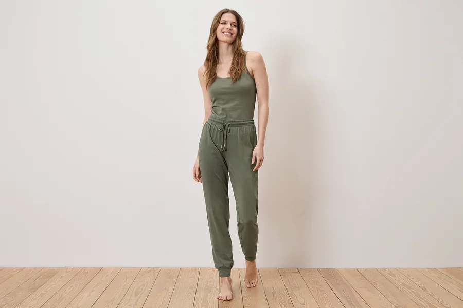 Eine Frau steht barfuß auf einem Holzboden und trägt ein olivgrünes Tanktop und eine Jogginghose.