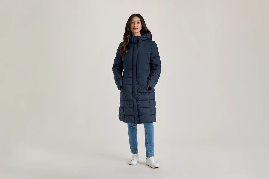 Frau steht, trägt einen langen, marineblauen, wattierten Mantel mit Kapuze, hellblaue Jeans und weiße Turnschuhe, die Hände in den Taschen.